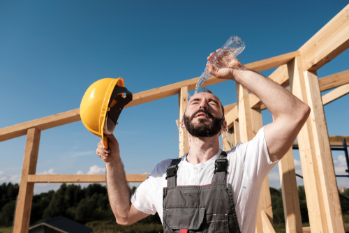 The man builder sits on the edge of the roof of the frame house, in a yellow helmet and gray overalls and a white T-shirt.