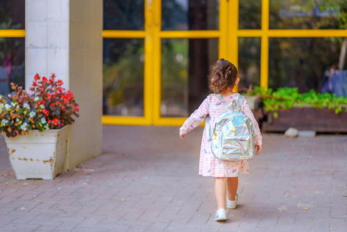 Curly cute little toddler girl back to school with holographic schoolbag or satchel, child going to kindergarten. 